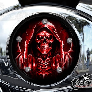 May include: A motorcycle engine cover with a red skull graphic. The skull is wearing a hood and giving the middle finger with both hands. The letters "F" and "U" are visible on the fingers. The cover is surrounded by a chrome housing.