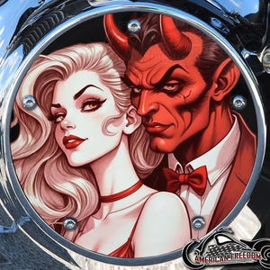 Puede incluir: Una pieza circular de motocicleta con una ilustración estilizada de una mujer y un diablo rojo. La mujer tiene cabello rubio y lápiz labial rojo, y el diablo tiene piel roja y cuernos. La imagen está enmarcada por un borde cromado, con el texto "AMERICAN FREEDOM" en la parte inferior.