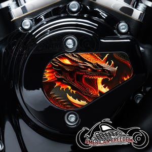 May include: Black motorcycle engine cover with a vibrant, detailed illustration of a red and orange dragon. The dragon's head is centered, with its mouth open, revealing sharp teeth. The cover has a glossy finish and is adorned with the text "AMERICAN FREEDOM".