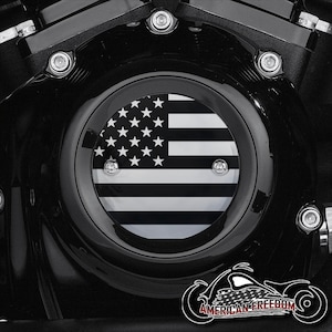 Puede incluir: Cubierta de motor de motocicleta negra con un emblema circular con un diseño de bandera estadounidense en blanco y negro. El emblema está asegurado con pernos plateados. Las palabras "AMERICAN FREEDOM" se muestran debajo.