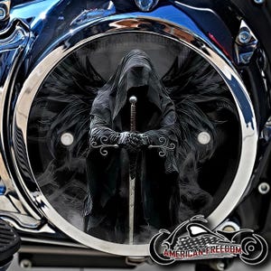 May include: A motorcycle engine cover with a detailed graphic of a cloaked figure with a sword and wings. The design is set within a chrome ring, with the words "AMERICAN FREEDOM" and a motorcycle logo at the bottom.