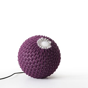K&ouml;nnte beinhalten: Ein lilafarbener, gestrickter Lampenschirm mit einem wei&szlig;en Zentrum und einem Kabel.