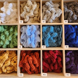 May include: Twelve samples of crocheted fabric in different colors, displayed in a wooden box. The colors include white, beige, gray, green, blue, red, purple, and yellow.