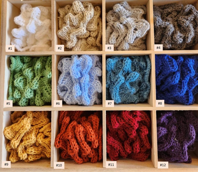 May include: Twelve samples of crocheted fabric in different colors, displayed in a wooden box. The colors include white, beige, gray, green, blue, red, purple, and yellow.