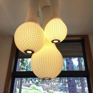 May include: A white, woven, pendant light fixture with three bulbs. The bulbs are shaped like teardrops and have a textured surface. The light fixture is hanging from the ceiling and is illuminated.