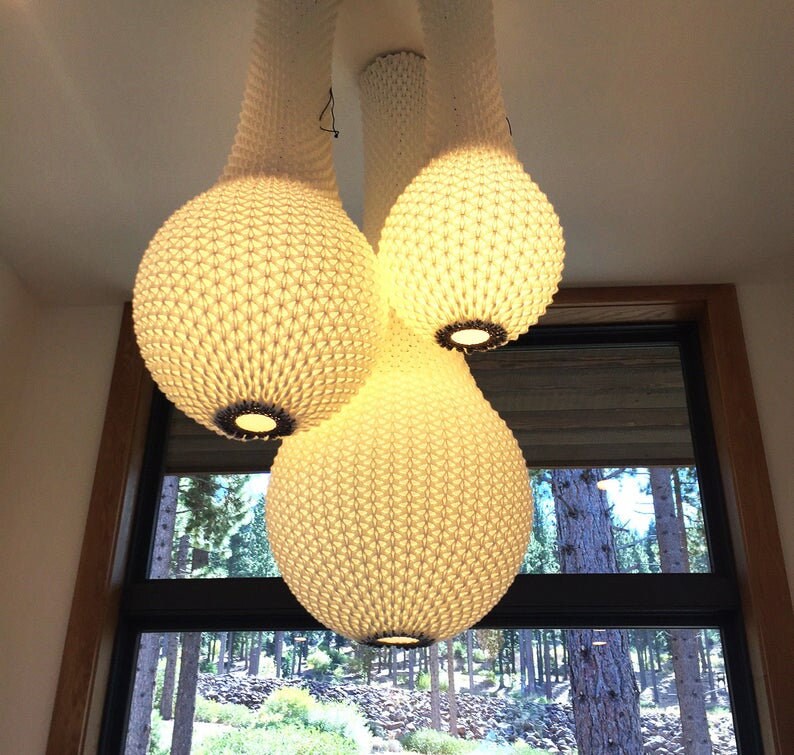 May include: A white, woven, pendant light fixture with three bulbs. The bulbs are shaped like teardrops and have a textured surface. The light fixture is hanging from the ceiling and is illuminated.