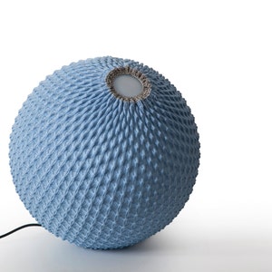 Op de afbeelding: Een blauwe gebreide lampenkap met een textuur en een kleine opening aan de bovenkant. De lampenkap is rond en heeft een snoer eraan bevestigd.