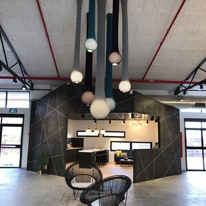 Puede incluir: Un espacio de oficina moderno con una lámpara única hecha de tejido tejido que cuelga del techo. La lámpara está compuesta por varias hebras largas y delgadas de tejido que se tejen juntas para crear una serie de orbes colgantes. Los orbes son en tonos de azul, gris y negro. El espacio está decorado con patrones geométricos en blanco y negro en las paredes e incluye dos sillas de mimbre negras con cojines blancos.
