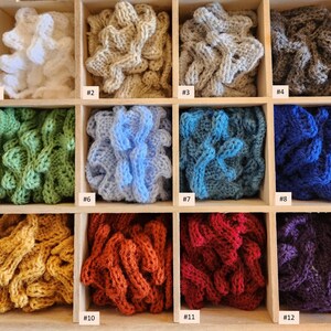 May include: Twelve samples of crocheted fabric in different colours, displayed in a wooden box. The colours include white, beige, grey, green, blue, red, purple, and yellow.
