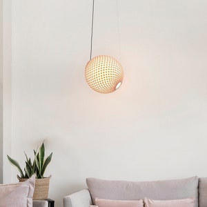 May include: A round, woven pendant light fixture hangs from a black cord. The light fixture is made of natural materials and has a light brown color. The light fixture is hanging in a room with white walls and a light brown couch.