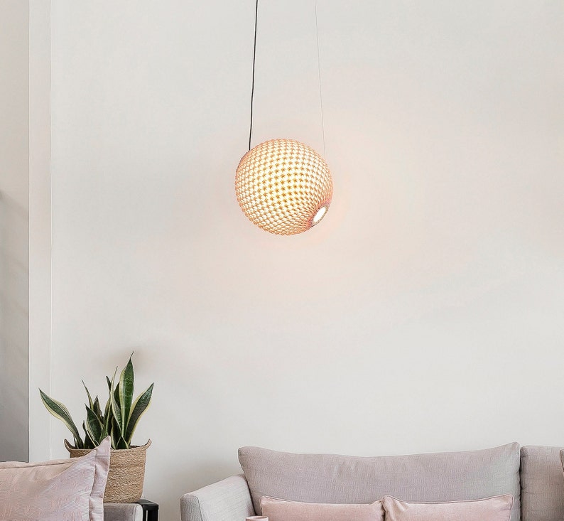 May include: A round, woven pendant light fixture hangs from a black cord. The light fixture is made of natural materials and has a light brown color. The light fixture is hanging in a room with white walls and a light brown couch.