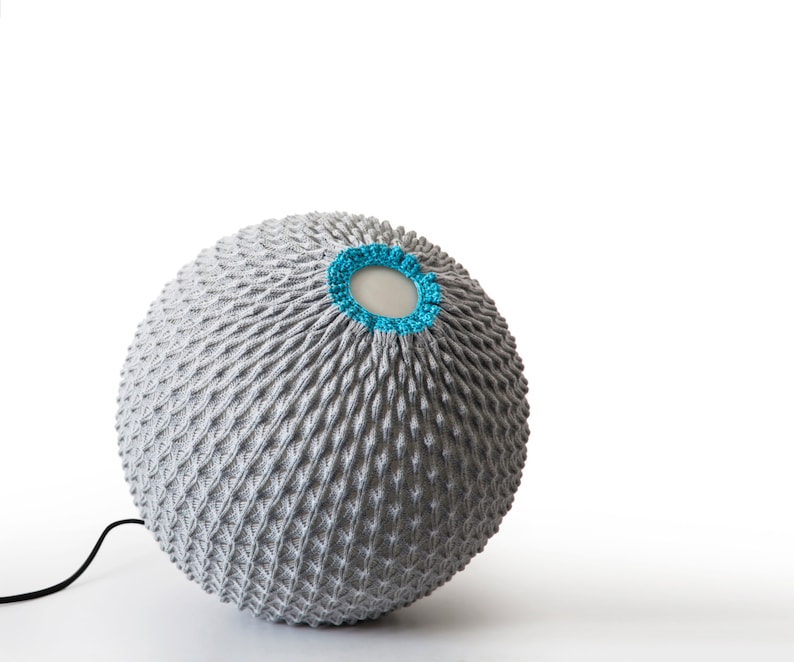 K&ouml;nnte beinhalten: Ein grauer, gestrickter Lampenschirm mit einem blauen Besatz um die obere &Ouml;ffnung. Der Lampenschirm ist rund und hat eine strukturierte Oberfl&auml;che.