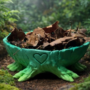 May include: A green, tree-stump-shaped bowl filled with dried brown leaves. The bowl has a heart-shaped cutout on the front and is sitting on a bed of dirt and moss. The bowl is a decorative item.