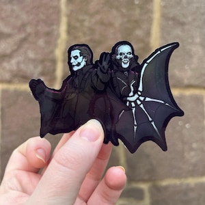 May include: A black sticker featuring two figures in dark attire with white skull-like faces. One figure has a bat-wing-shaped accessory. The sticker is held by a hand against a textured brown background.