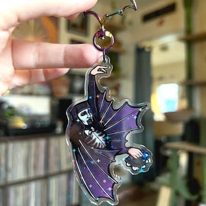 May include: A clear acrylic keychain featuring a character in a dark cloak with purple bat wings, holding a tambourine. The keychain has a rainbow-colored bat-shaped clasp and a silver ring.