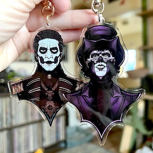 Könnte beinhalten: Zwei Schlüsselanhänger aus Acryl mit detaillierten Illustrationen von Charakteren mit aufwendigem Make-up und Kleidung. Ein Schlüsselanhänger hat einen Charakter mit weißer Gesichtsbemalung und dunkler Uniform. Der andere hat einen Charakter in einem lila Hut und Outfit.