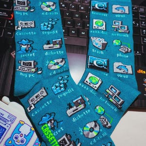 Puede incluir: Un par de calcetines de color verde azulado con un patrón repetitivo de arte de píxeles de iconos informáticos antiguos y etiquetas que incluyen my pc, cd-rom, cassette, regedit, secret, diskette, browser, virus, aaaa.doc, network, trash y phone.