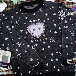 Puede incluir: Jersey negro de punto con un diseño de cara de gato en forma de corazón, estrellas y texto japonés. El jersey presenta detalles en rosa y blanco, cuello, puños y dobladillo de canalé. La palabra "Cassette" está impresa en la manga derecha.