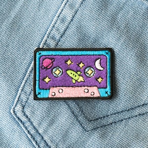 May include: A blue and pink embroidered patch shaped like a cassette tape with a purple background. The patch features a rocket ship, stars, a crescent moon, and a planet.