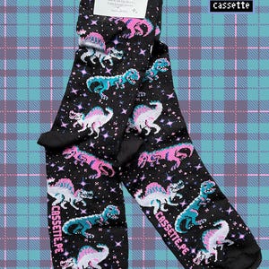 Puede incluir: Calcetines negros con un patrón repetido de dinosaurios rosas y turquesas y estrellas. Los calcetines tienen el texto "cassette.pe" impreso en el lateral. Una etiqueta está adherida a los calcetines.