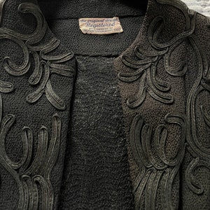 May include: Black jacket with a black embroidered design. The jacket has a label that reads "an original design Registered by member of FASHION ORIGINATORS GUILD".