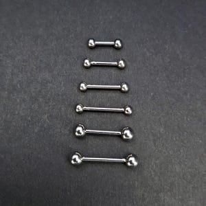 Puede incluir: Una colección de joyas para el cuerpo de estilo barra con mancuernas de color plateado. Cada pieza presenta una barra recta con una bola redondeada en cada extremo. Las joyas están dispuestas verticalmente sobre un fondo gris oscuro.