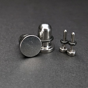 May include: A selection of stainless steel body jewellery. The image shows two flat-faced plugs, a rounded plug with a black rubber ring, and two small barbell-style pieces. The items are on a dark grey surface.