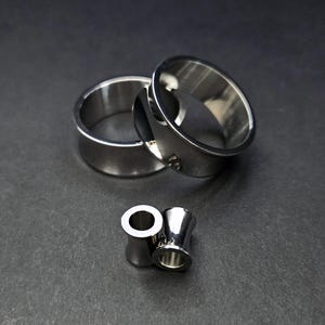 Könnte beinhalten: Ein Set silberfarbener Ohr-Gauges in verschiedenen Größen. Die Gauges bestehen aus glänzendem Metall und haben eine glatte, polierte Oberfläche. Das Bild zeigt zwei große und zwei kleinere Gauges.