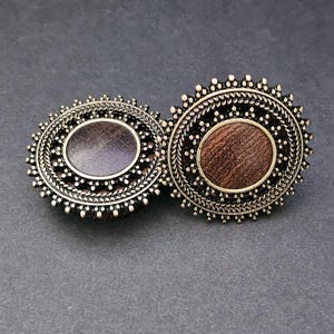 May include: A pair of silver-coloured, circular earrings with a detailed, beaded border. Each earring has a central, circular inlay, one in a dark purple hue and the other in a warm brown. The earrings are set against a dark grey background.