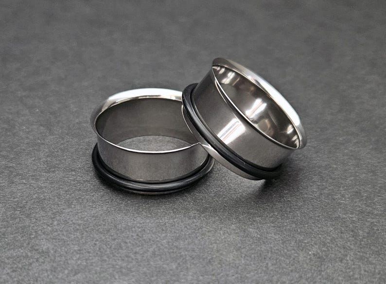 May include: Two polished, silver-colored ear gauges with black rubber O-rings. The tunnels are circular and have a smooth, reflective surface. They are displayed on a dark gray surface.