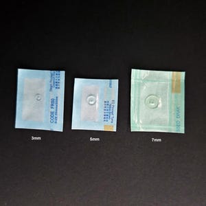 Op de afbeelding: Drie kleine, verzegelde medische pleisters van verschillende afmetingen, 3 mm, 5 mm en 7 mm, worden getoond. Elke pleister heeft een helder, cirkelvormig onderdeel dat zichtbaar is door de verpakking. Tekst is zichtbaar op de verpakking.