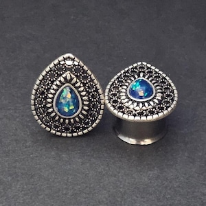 Könnte beinhalten: Ein Paar silberne tropfenförmige Ohrdehner mit einem schwarzen Filigranmuster und einem schillernden blauen Opal-Stein in der Mitte.