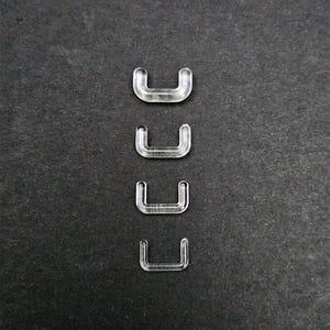 Puede incluir: Cuatro conectores de plástico transparente en forma de U están dispuestos verticalmente sobre un fondo gris oscuro. Los conectores están hechos de un material transparente, lo que permite ver el fondo a través de ellos. Las formas son idénticas.