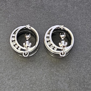 May include: Two silver-toned ear gauges featuring a crescent moon design with a sitting cat silhouette. The moon is adorned with small, clear crystals and a star. The gauges are set against a gray background.