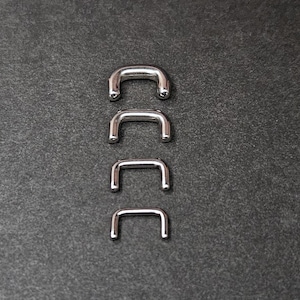 Puede incluir: Cuatro piezas de ferretería metálicas plateadas en forma de U, de diferentes tamaños, están dispuestas verticalmente sobre un fondo gris oscuro. Las piezas tienen una superficie lisa y reflectante y bordes redondeados.