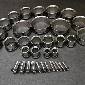 May include: A collection of stainless steel ear gauges in various sizes, arranged on a dark gray surface. The gauges include tunnel and plug styles, showcasing a range of diameters for body modification. The metal has a polished, reflective finish.