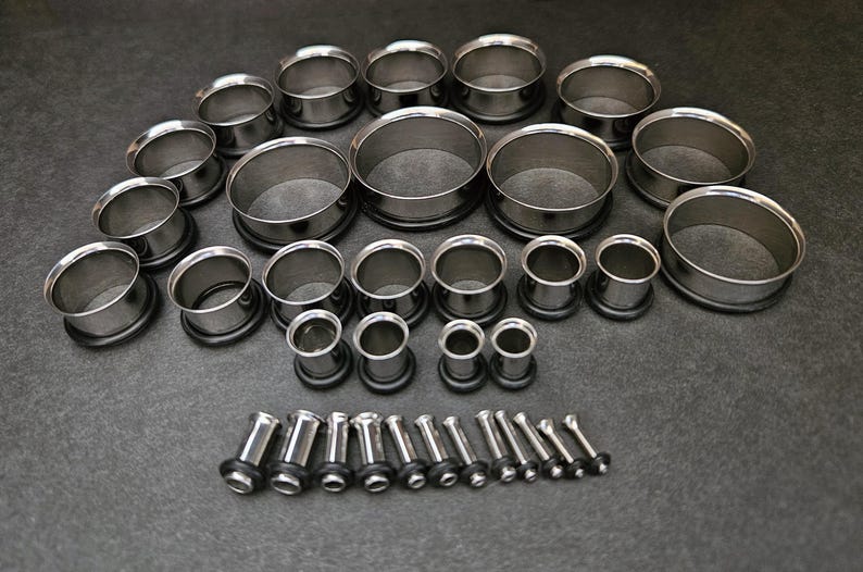 May include: A collection of stainless steel ear gauges in various sizes, arranged on a dark gray surface. The gauges include tunnel and plug styles, showcasing a range of diameters for body modification. The metal has a polished, reflective finish.