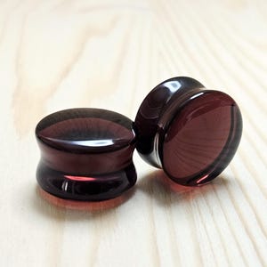 May include: Two dark purple glass ear gauges, one slightly larger than the other, on a light wood surface.