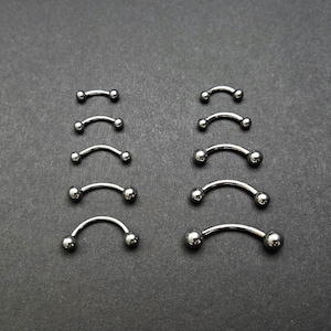 May include: An assortment of curved barbell piercings in different sizes. Each piece has a curved, silver-coloured bar with a small ball at each end. The piercings are arranged in two columns against a dark grey background.
