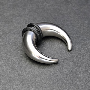 Può includere: Un dilatatore per orecchio lucido, color argento, con un design curvo a forma di corno. Il dilatatore è realizzato in metallo e ha una superficie liscia e riflettente. Un anello di gomma nero è attaccato. L'articolo è esposto su una superficie grigio scuro.