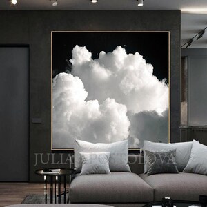 Large framed artwork featuring a black and white cloudscape. The image showcases fluffy white clouds against a dark background, creating a dramatic contrast. The artwork is displayed in a modern living space with a gray sofa and pillows.