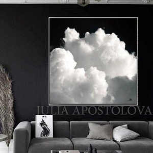 A large, framed black and white cloudscape art piece hangs above a gray sofa. The artwork features a dramatic depiction of fluffy white clouds against a dark sky. The room is decorated in shades of gray and black, with a modern aesthetic.