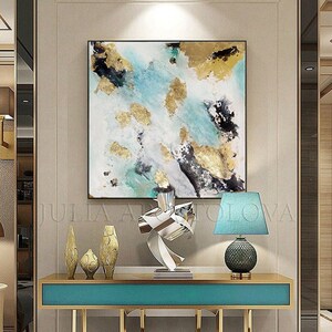 May include: Abstract painting with gold leaf accents on a white canvas, displayed above a teal console table with gold legs. The table is decorated with a silver metal sculpture and three gold ceramic vases.