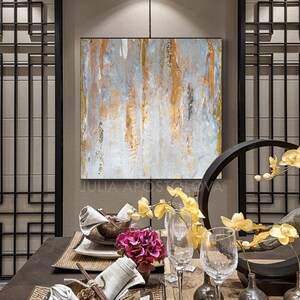 May include: Abstract painting with gold and gray vertical brushstrokes. The artwork is framed in a gold border and hangs above a dining table set with glassware, flowers, and decorative elements. The artist's name, Julia Apostolova, is visible at the bottom.