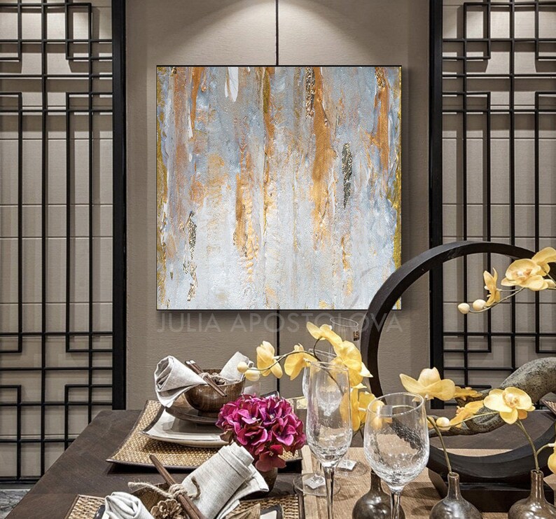 May include: Abstract painting with gold and gray vertical brushstrokes. The artwork is framed in a gold border and hangs above a dining table set with glassware, flowers, and decorative elements. The artist's name, Julia Apostolova, is visible at the bottom.