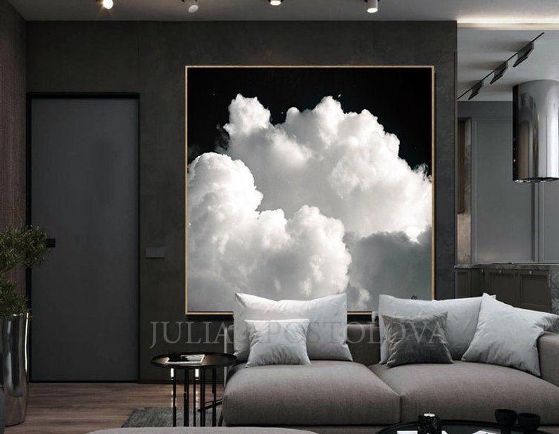 Large framed artwork featuring a black and white cloudscape. The image showcases fluffy white clouds against a dark background, creating a dramatic contrast. The artwork is displayed in a modern living space with a gray sofa and pillows.