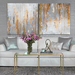 May include: Abstract art diptych with vertical brushstrokes in gold, white, and gray. The artwork hangs above a white sofa with silver pillows, a glass coffee table, and a vase of pink flowers. The room has a modern, elegant aesthetic.
