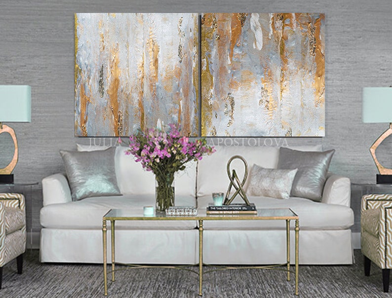 May include: Abstract art diptych with vertical brushstrokes in gold, white, and gray. The artwork hangs above a white sofa with silver pillows, a glass coffee table, and a vase of pink flowers. The room has a modern, elegant aesthetic.