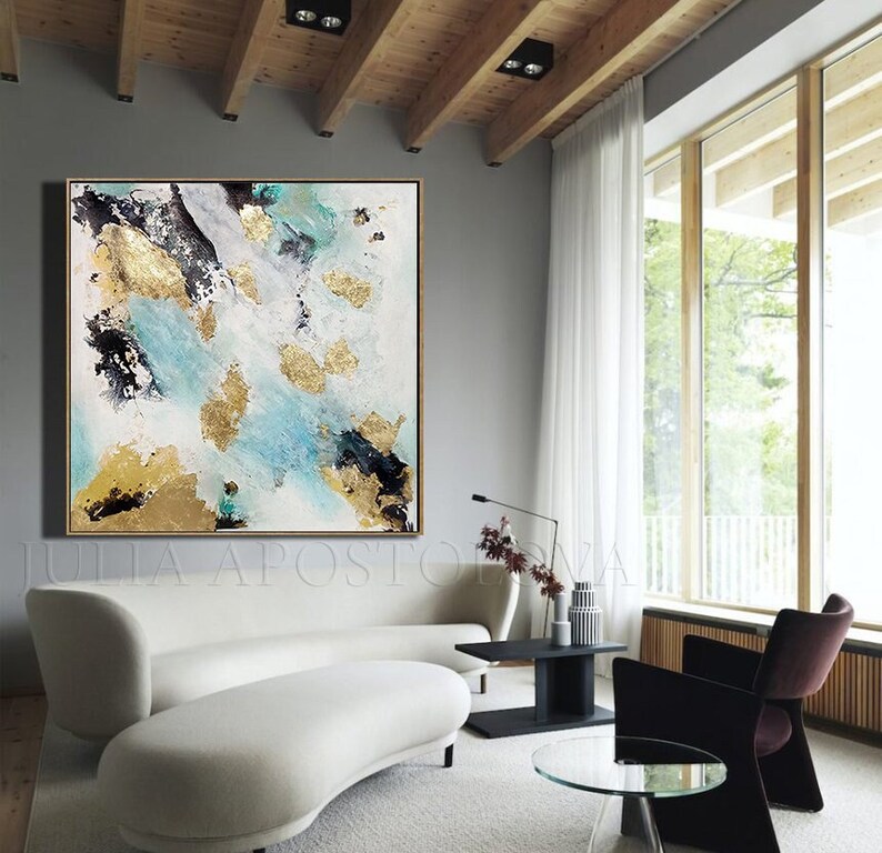 May include: Abstract painting with gold leaf accents on a white canvas. The painting features shades of blue, black, and white. The painting is hanging on a gray wall in a modern living room.