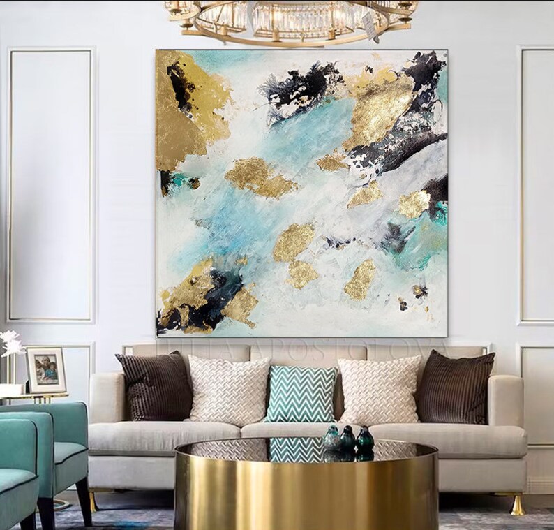 May include: Abstract painting with blue, white, and gold accents. The painting features a modern, abstract design with splashes of color and gold leaf. The painting is framed and hanging on a white wall above a couch.